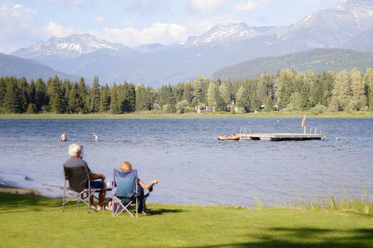 An older man and woman sit in folding chairs on a grassy shore looking out at a blue lake and floating dock with trees and mountains in the distance